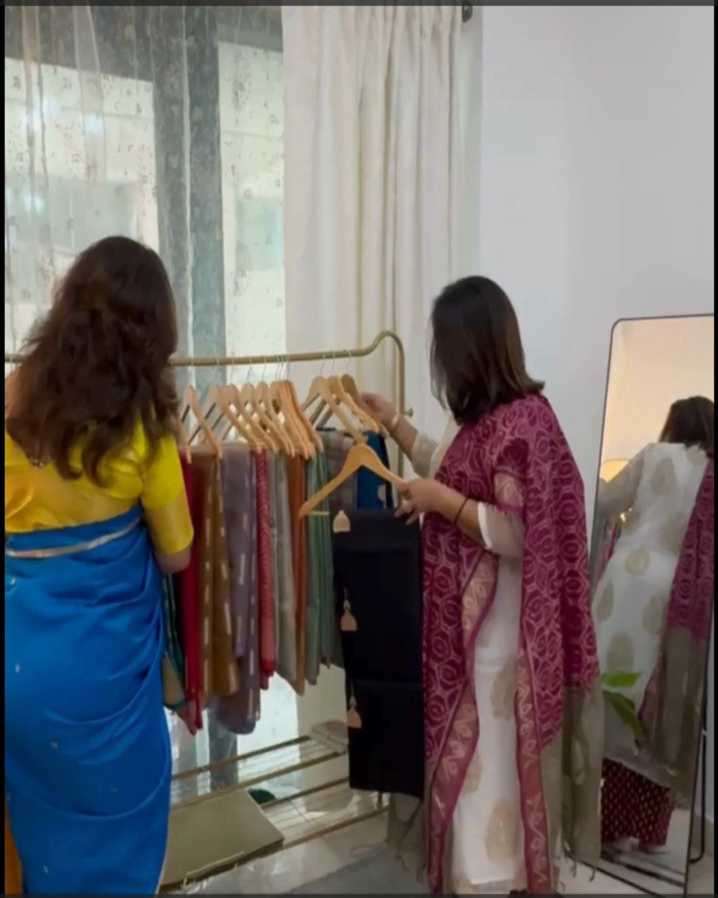 Two women browsing a gold clothing rack filled with colorful garments in an indoor boutique setting. One woman wears a bright blue saree with a yellow top, and the other wears a white kurta with a pink patterned shawl.